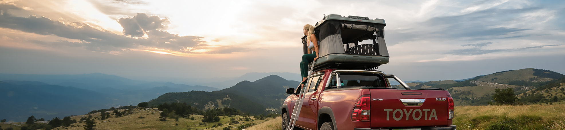 Toyota Hilux rossa equipaggiata con una tenda da tetto James Baroud grand Raid EVO e una donna seduta all'uscita della tenda.